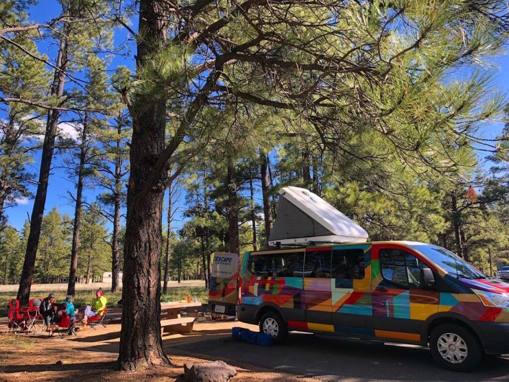 Campers sit near a van in the forest.