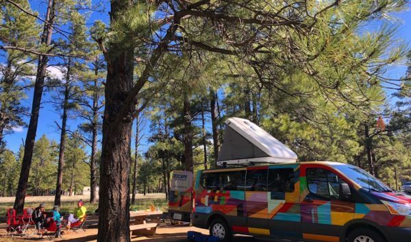 Campers sit near a van in the forest.